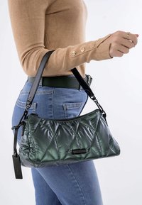 Doorstikte donkergroene handtas met een glanzende afwerking, met een zwarte strap en een label, vastgehouden tegen een casual denimoutfit.