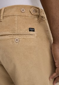 Pantaloni in velluto a coste color beige chiaro con finitura strutturata, dotati di una tasca posteriore con etichetta logo e due bottoni vicino alla vita.