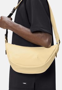 Borsa a tracolla gialla con una texture liscia e forma curvilinea, dotata di una tracolla nera regolabile e dettagli in metallo dorato.