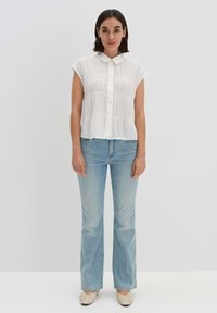 White short-sleeve shirt with textured fabric and button closure. Paired with light blue flared jeans and beige slip-on shoes.