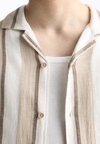 Camicia di lino con righe verticali beige e marroni, colletto abbottonato e un tessuto leggermente strutturato. Sotto, una canotta bianca a costine.