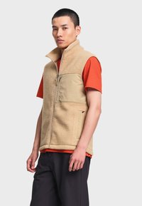 Homme portant une veste polaire beige par-dessus une chemise orange à manches courtes et un pantalon noir, debout devant un fond clair uni.