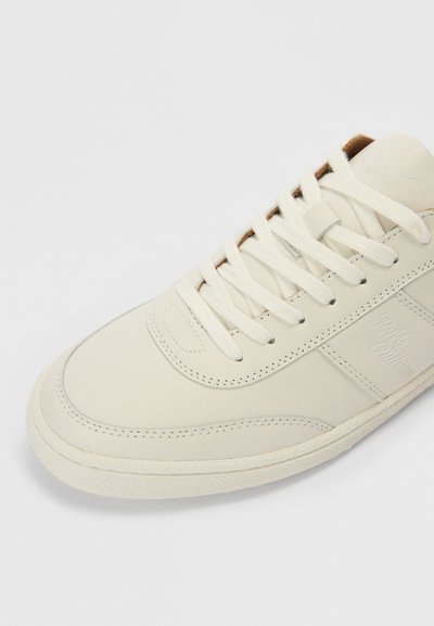 Balts ādas sporta apavs ar apaļu purngalu, plānām auklām un fakturētu gumijas zoli. Uz tā ir redzamas šuvju līnijas un sānos iespiests logo.