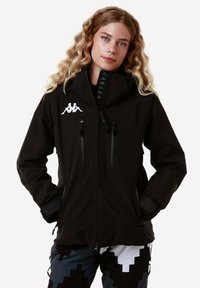 Giacca nera impermeabile con cappuccio, dotata di due tasche frontali con zip, logo bianco sul petto e fodera interna texturizzata.