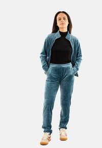 Femme debout avec les mains dans les poches, portant un survêtement en velours teal, un t-shirt noir et des baskets beiges avec des accents bordeaux.