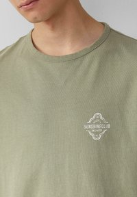 Oliwkowa bawełniana koszulka z okrągłym dekoltem i małym białym logo "SUNSHINECLUB" na piersi. Miękka faktura, casualowy styl.