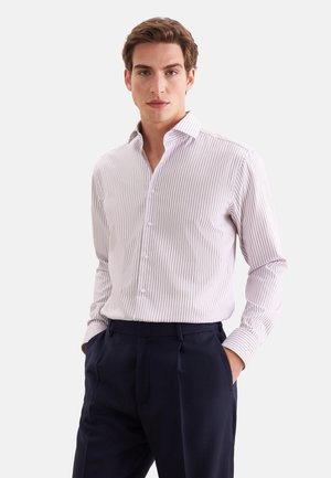 Jeune homme portant une chemise à rayures rose clair et blanc avec un pantalon bleu marine foncé, les mains dans les poches, debout devant un fond uni.