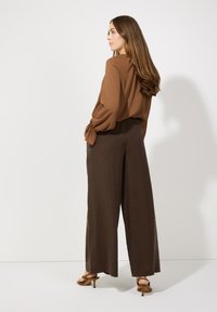 Braune Bluse mit geschwollenen Ärmeln, kombiniert mit hoch taillierten, weiten braunen Hosen. Das Modell trägt braune Sandalen mit Absatz.