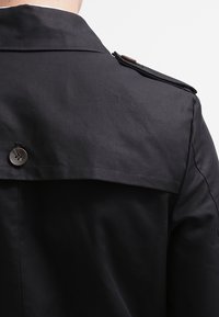 Czarny płaszcz trench z wyprofilowanym kołnierzem, zapięciem na epauletki oraz gładką fakturą. Posiada zapięcie na guziki z tyłu.