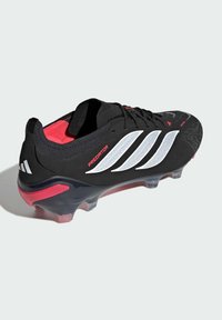 Μαύρο ποδοσφαιρικό παπούτσι με υφασμένο επάνω μέρος, κόκκινες λεπτομέρειες, τρεις λευκές διαγώνιες ρίγες και διάφανη σόλα με ροζ απόχρωση.