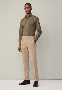 Olijfgroene button-up shirt met kraag, gecombineerd met beige, op maat gemaakte broeken. Kenmerkend voor het strakke ontwerp en bruine leren dresschoenen.
