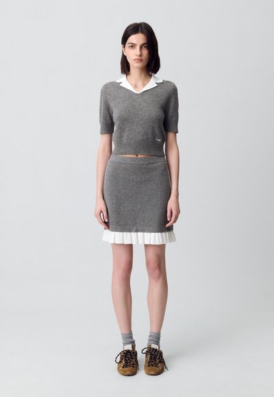Suéter gris corto con cuello blanco, combinado con una falda gris con un dobladillo blanco con volantes. La modelo lleva zapatos beige y calcetines grises.