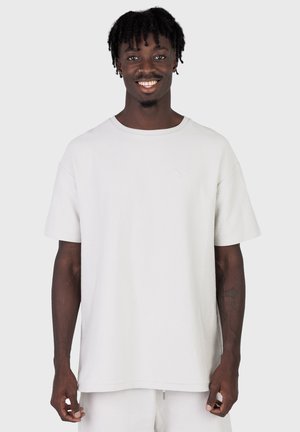 Lachende man met korte dreadlocks, gekleed in een los wit kort mouw shirt en bijpassende witte broek tegen een effen achtergrond.