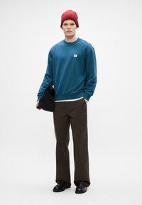 Felpa teal con collo a giro e patch del logo, abbinata a pantaloni verde oliva. Completata con un berretto a maglia rosso e una borsa nera.