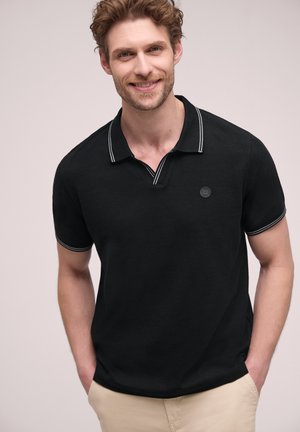 Smilende mand med krøllet hår iført sort polo med hvide kanter og beige bukser, stående med hænderne i lommerne.