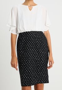 Blusa bianca con scollatura a forma di chiave e maniche a soffietto, abbinata a una gonna a tubino nera con pois bianchi. Texture morbide.