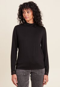 Schwarzes Langarmoberteil mit einem hohen Hals und Spitzenakzenten an den Ärmelbündchen, aus glattem Stoff gefertigt. Kombiniert mit grauen Jeans.