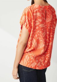 Blusa de manga corta, de color naranja brillante, con tela texturizada, que presenta un patrón floral y un detalle de botón en la parte trasera.