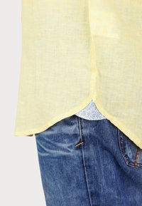 Camicia di lino gialla con un orlo curvo, indossata sopra una camicia a fantasia. Indossata con jeans di denim blu caratterizzati da una cucitura visibile.
