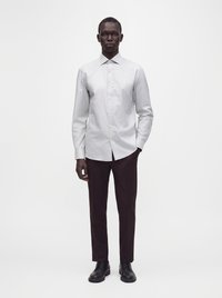 Homme debout face à l'avant portant une chemise gris clair boutonnée, un pantalon foncé et des chaussures de ville noires sur un fond blanc uni.