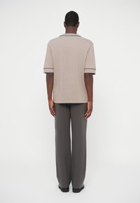 Polo shirt beige a maniche corte con bordi neri, vestibilità comoda, abbinato a pantaloni grigi a gamba larga e scarpe nere, mostrato da dietro.