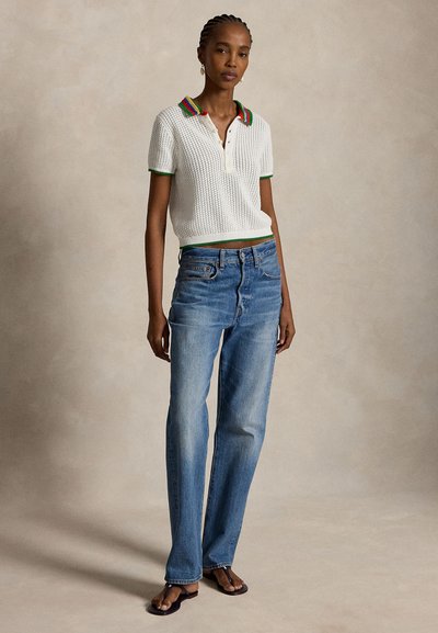 Ψηλή γυναίκα με πλεξούδες φοράει λευκή πλεκτή μπλούζα πόλο με πολύχρωμο γιακά, μπλε τζιν με ίσια μπατζάκια και μαύρα σανδάλια, όρθια σε μπεζ πάτωμα.