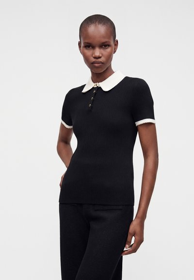 Femme portant un top noir ajusté à manches courtes avec un col blanc et une patte de boutonnage, debout devant un fond clair uni.