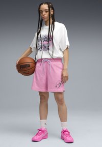 Persoon met lange vlechten, gekleed in een wit grafisch T-shirt, roze mesh shorts en roze sneakers, met een basketball in één hand.