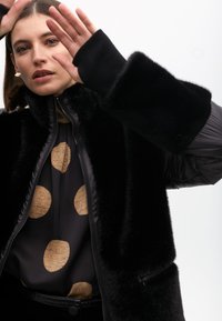 Black faux fur jacket with shiny sleeves, worn over a dark blouse featuring large golden polka dots; model poses with hands raised.