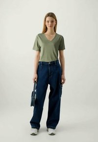 T-shirt verde-oliva com decote em V, jeans azuis folgados e sapatilhas brancas. O modelo segura uma pequena mala azul. Design simples com um ajuste casual.