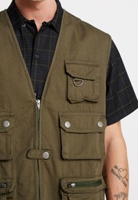 Olivgrüner Weste mit mehreren Taschen, Reißverschluss vorne und Druckknopfverschlüssen. Aus robustem Stoff gefertigt, mit einem Ringdetail und strukturiertem Finish.