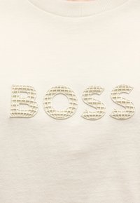 Υφασμα εκρού χρώματος με ανάγλυφα, υφασμένα γράμματα που σχηματίζουν τη λέξη "BOSS" σε μοτίβο πλέγματος σε ένδυμα με στρογγυλή λαιμόκοψη.