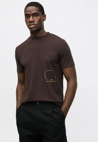 T-shirt de algodão castanho com decote arredondado. Apresenta um gráfico amarelo de um rosto no lado esquerdo do peito. Combinado com calças pretas.