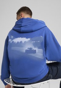 Plava dukserica s grafikom trkaćeg automobila na poleđini, s glatkom teksturom i opuštenim krojem.