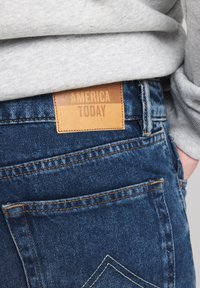 Blue denim jeans with a smooth texture, featuring a leather label reading "AMERICA TODAY" near the back pocket. Stitching in contrasting orange.