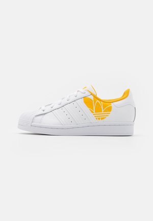Biały sneaker Adidas z trzema paskami i żółtym logo Adidas po boku, żółta wyściółka oraz biała gumowa podeszwa.