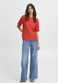 Rød top med puffede ærmer og struktureret mønster, parret med brede blå denimjeans. Sættet har en afslappet pasform og casual stil.