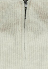 Off-white strikkegenser med frontglidelås. Teksturert ribbestrikkedetalj gjennom hele genseren med en subtil glans. Glidelåsen er av metall med en pull-tab.