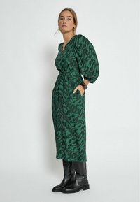 Vihreä kuvioitu mekko, jossa on kietaisusuunnittelu, poimutettu pinta ja sivutaskut. Yhdistetty mustiin polvipitkiin saappaisiin, joissa on paksu pohja.