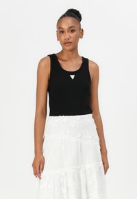 Svart ribbad tank top med rund ringning och en triangulär silvertag i mitten; matchad med en vit broderad lager-på-lager kjol.