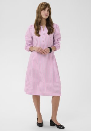 Žena s dlhými hnedými vlasmi v ružovo-bielych pruhovaných kolenných šatách a čiernych nízkych podpätkoch, stojaca so zloženými rukami.