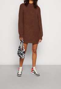 Pull surdimensionné marron en tricot avec manches longues, associé à un sac noir et blanc à motifs et des baskets grises avec des accents rouges.