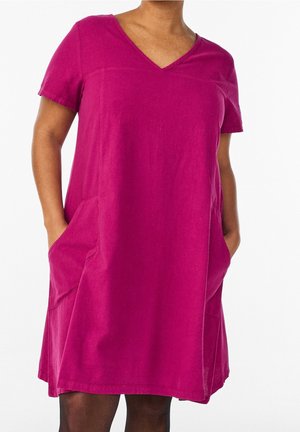 Femme portant une robe magenta à manches courtes avec col en V et poches, mains dans les poches, vue des épaules jusqu'au milieu des cuisses sur fond blanc.