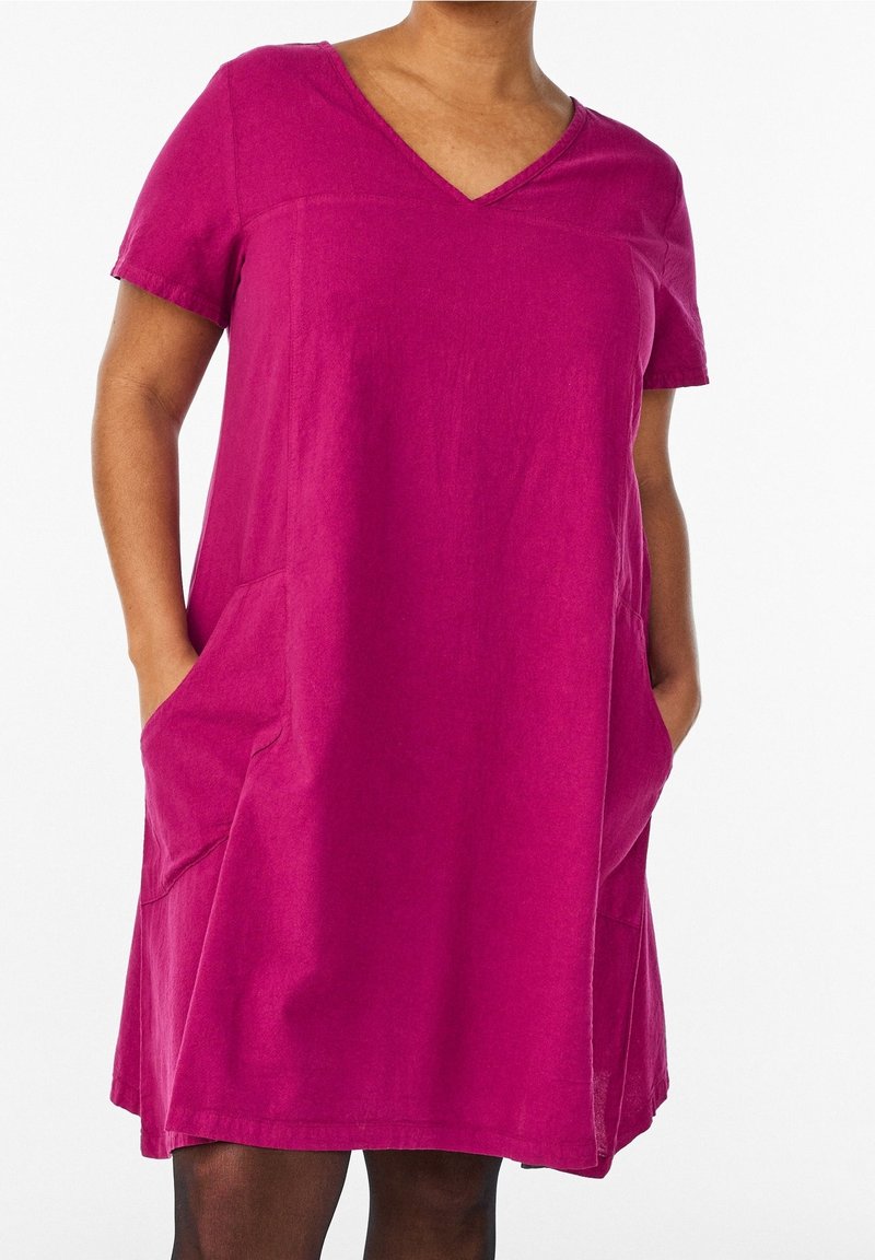 Femme portant une robe magenta à manches courtes avec col en V et poches, mains dans les poches, vue des épaules jusqu'au milieu des cuisses sur fond blanc.