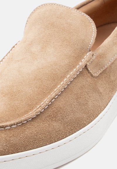 Γαλακτερό-καφέ σουέντ παπούτσι τύπου slip-on με λευκή ραφή και λευκή λαστιχένια σόλα, που δείχνει κοντινή λεπτομέρεια του πάνω μπροστινού και πλάγιου μέρους.