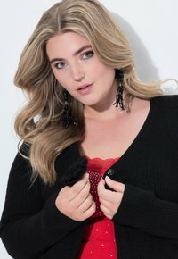 Blondýnka se zelenýma očima, která má na sobě černé korálkové náušnice s třásněmi a drží černý kardigan přes červený top s krajkou a kamínky.