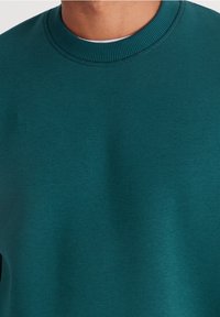 Bluza z okrągłym dekoltem w kolorze morskim, wykonana z miękkiego materiału, z żebrowanym wykończeniem przy szyi i gładką fakturą, bez widocznych wzorów ani akcentów.