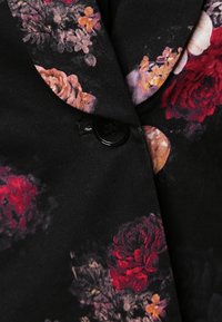 Tissu noir à motif floral avec des fleurs rouges, roses et jaunes entourant un bouton noir sous un col à revers.
