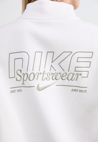 White sweatshirt featuring large green "NIKE" text, "Sportswear" in smaller script, and "EST. 1972" and "JUST DO IT" details. Soft texture.