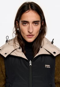 Giacca isolante nera e beige con colletto alto, chiusura con zip e logo ricamato. Tessuto liscio con finitura lucida.